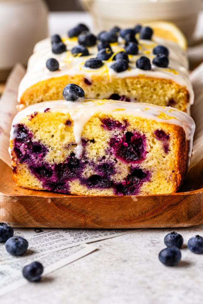 blueberry lemon loaf with lemon glaze on top, sliced on top of a wooden plate.