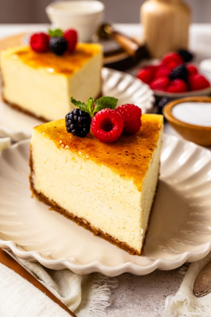 slice of Crème Brûlée Cheesecake on top of a plate, with berries on top of the cheesecake slice, and another slice of cheesecake on the back.