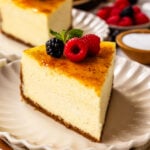 slice of Crème Brûlée Cheesecake on top of a plate, with berries on top of the cheesecake slice, and another slice of cheesecake on the back.