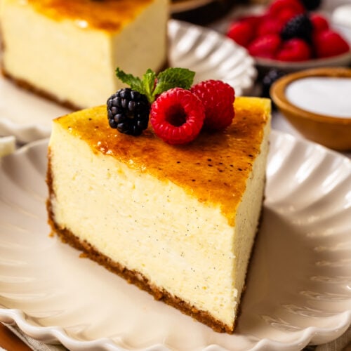 slice of Crème Brûlée Cheesecake on top of a plate, with berries on top of the cheesecake slice, and another slice of cheesecake on the back.
