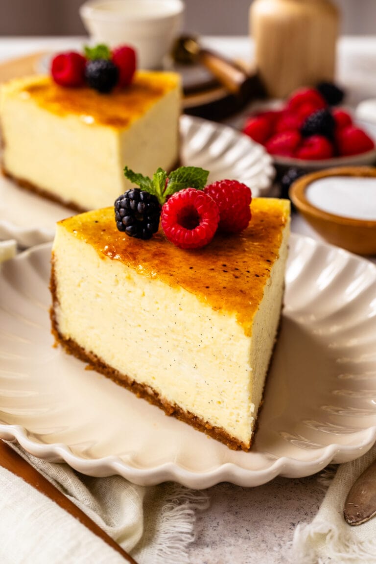 slice of Crème Brûlée Cheesecake on top of a plate, with berries on top of the cheesecake slice, and another slice of cheesecake on the back.