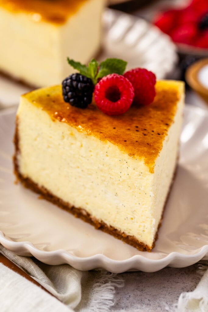 slice of Crème Brûlée Cheesecake on top of a plate, with berries on top of the cheesecake slice, and another slice of cheesecake on the back.