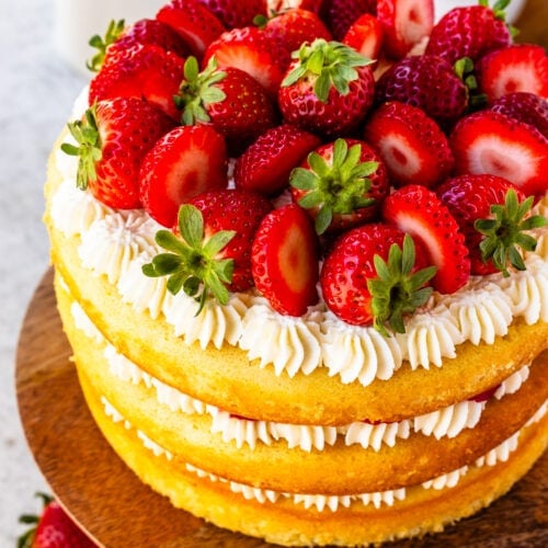 Strawberry Shortcake Cake, layers of vanilla cake with strawberries and whipped cream in between the layers and on top, on a wooden cake plate, with strawberries around.