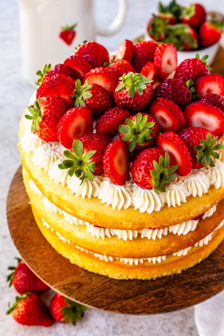 Strawberry Shortcake Cake, layers of vanilla cake with strawberries and whipped cream in between the layers and on top, on a wooden cake plate, with strawberries around.