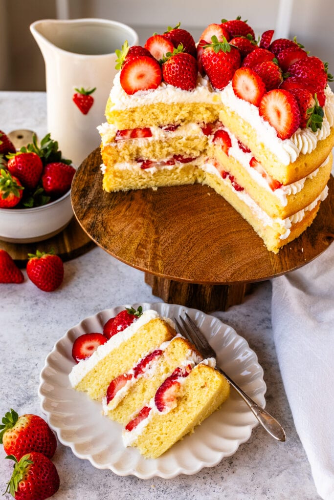 slice of strawberry cake on a plate, with whipped cream and fresh strawberries in the middle, and the cake sliced in the back, on a wooden cake plate.