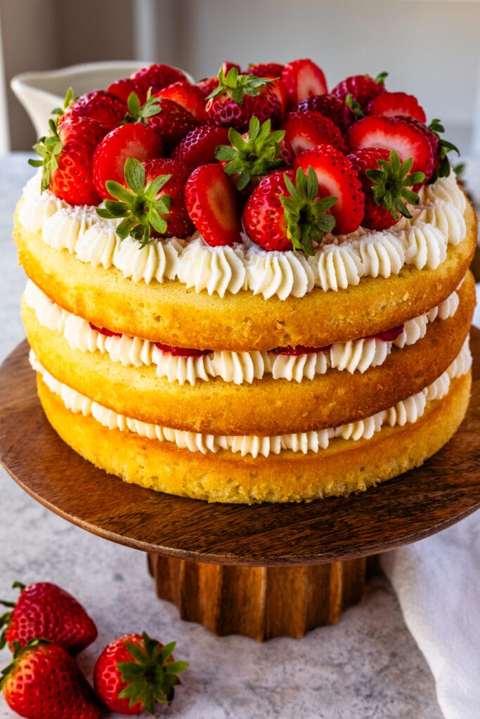 Strawberry Shortcake Cake, layers of vanilla cake with strawberries and whipped cream in between the layers and on top, on a wooden cake plate, with strawberries around.