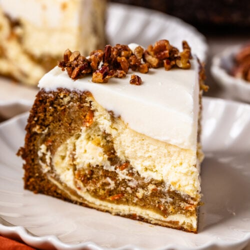 slice of carrot cake cheesecake on a plate, with cream cheese frosting and candied pecans on top.
