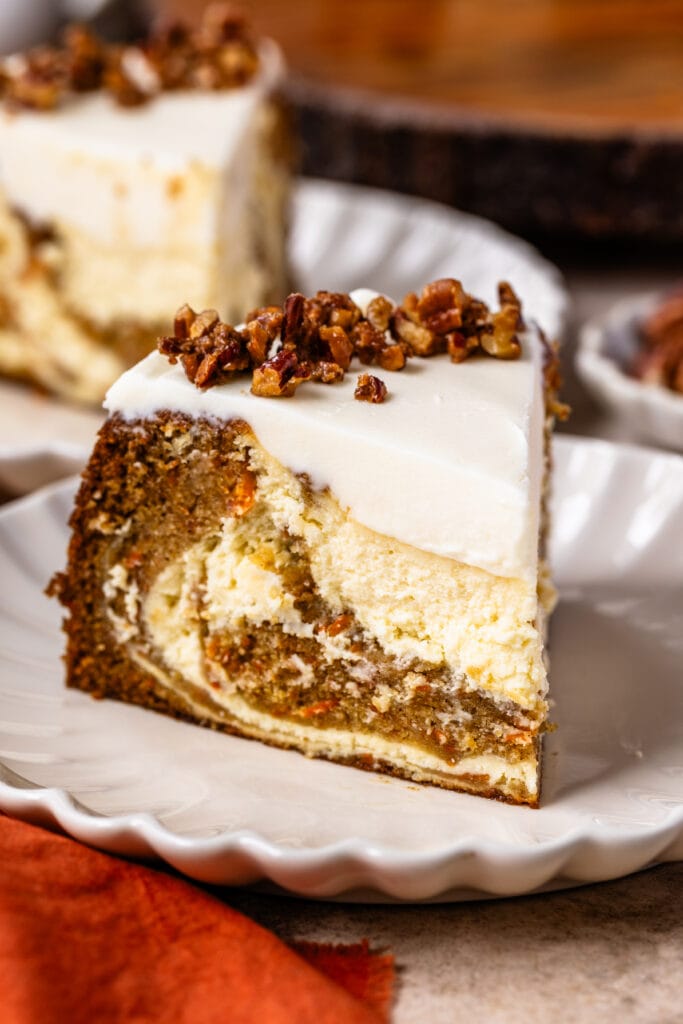 slice of carrot cake cheesecake on a plate, with cream cheese frosting and candied pecans on top.