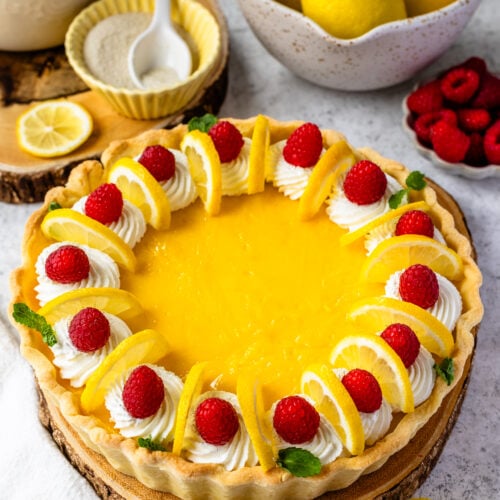 Lemon pie with raspberries and lemon slices on top, and mint leaves, on top of a wooden board.