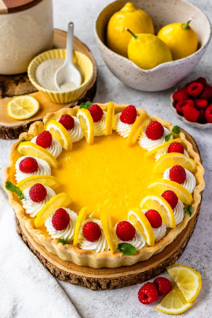 Lemon pie with raspberries and lemon slices on top, and mint leaves, on top of a wooden board.