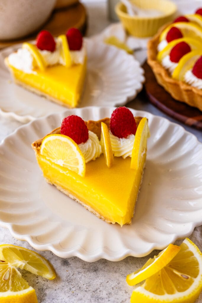 slice of lemon tart on a plate, with lemon slices on top and raspberries, and whipped cream.