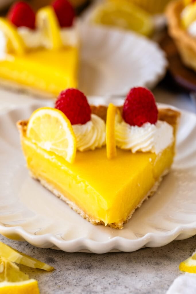 slice of lemon tart on a plate, with lemon slices on top and raspberries, and whipped cream.