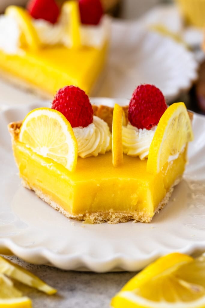 slice of lemon tart on a plate, with lemon slices on top and raspberries, and whipped cream.