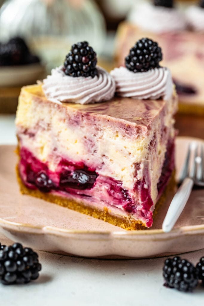 slice of blackberry cheesecake with graham cracker crust, and blackberry sauce mixed into the cheesecake batter, with whipped cream on top and blackberry.