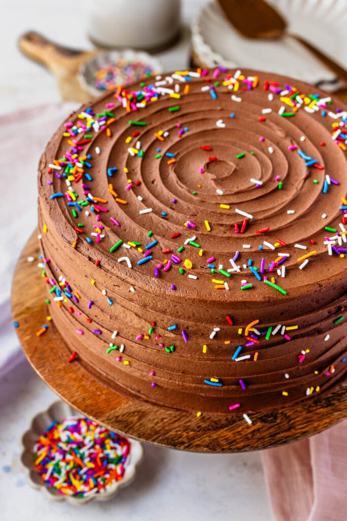 chocolate cake on top of a wooden cake stand with sprinkles on top of the cake and in a bowl on the bottom.