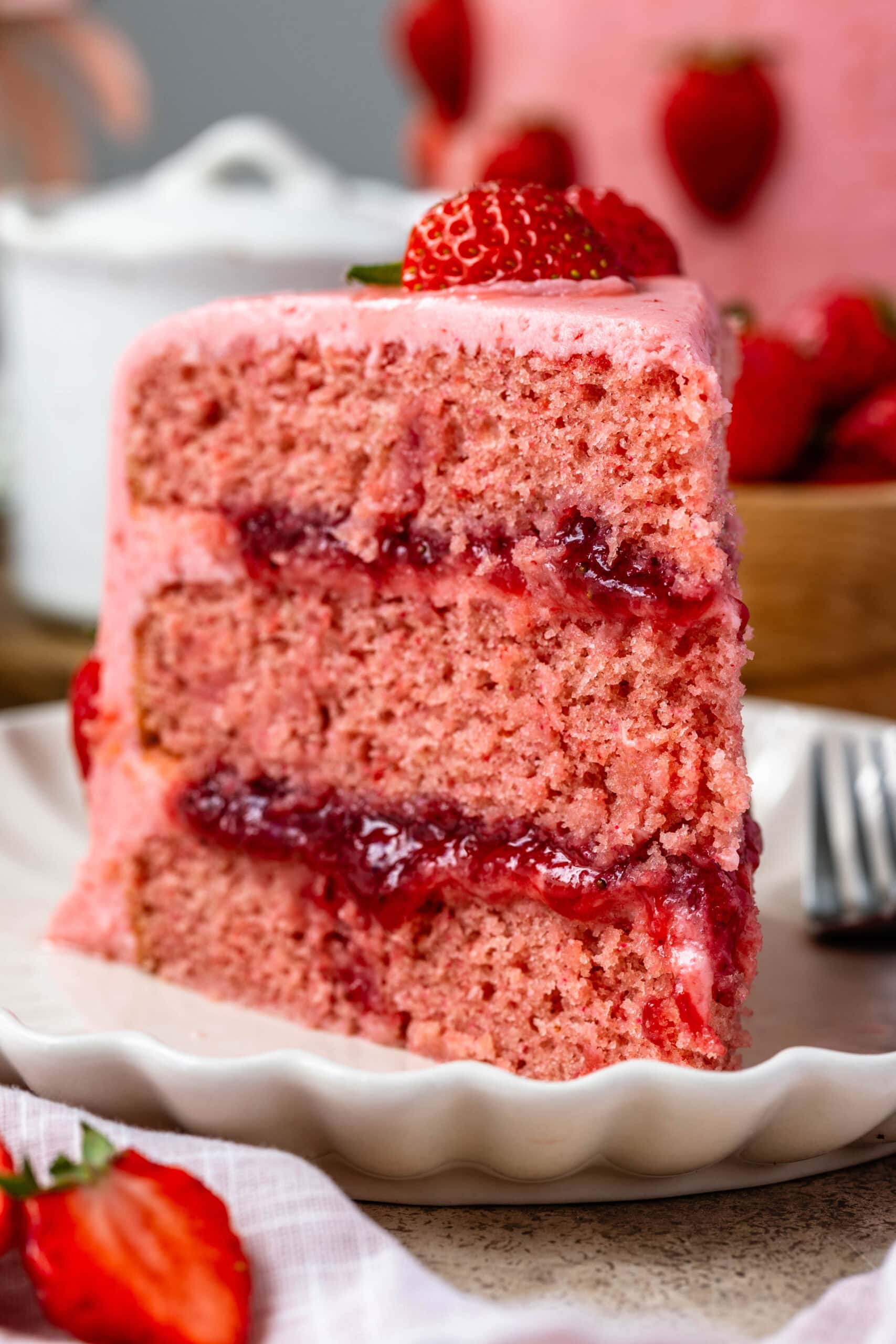 slice of strawberry cake on a plate, the cake is pink filled with strawberry jam and frosted with pink strawberry frosting.