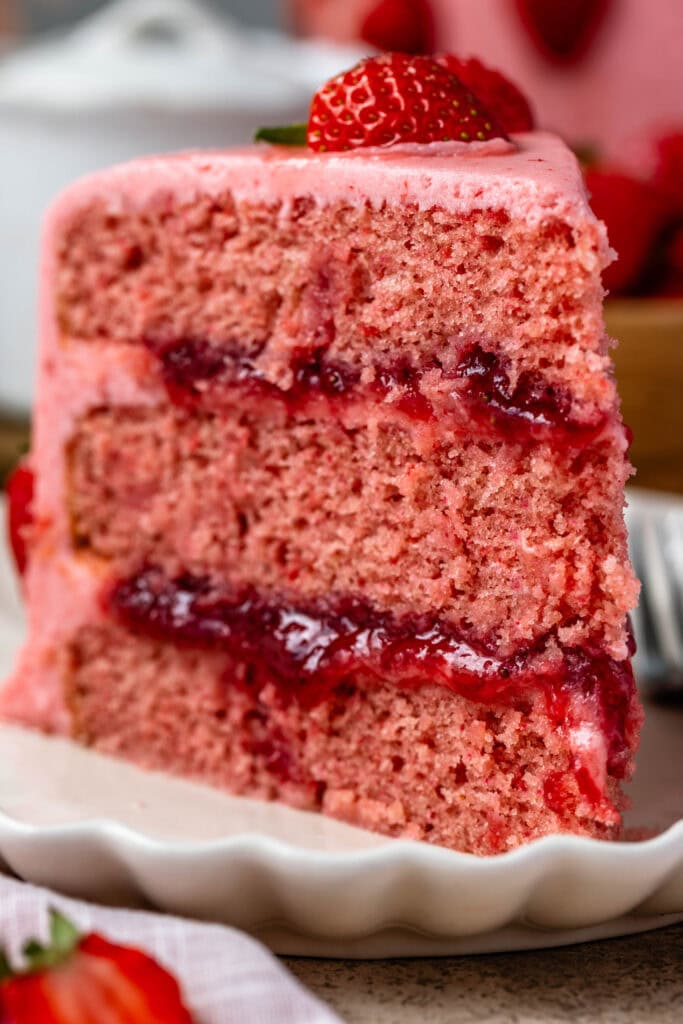 slice of strawberry cake on a plate, the cake is pink filled with strawberry jam and frosted with pink strawberry frosting.