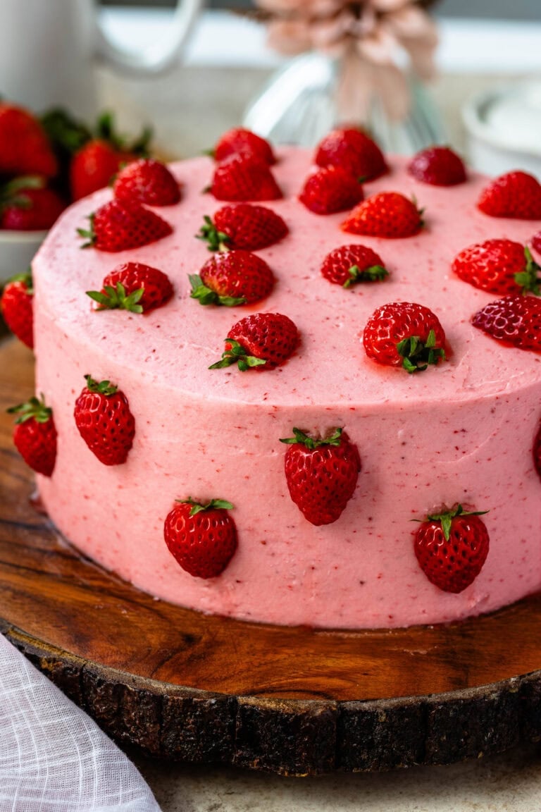 strawberry cake, a pink cake on top of a wooden board, the cake has pink frosting and is decorated with halved strawberries.