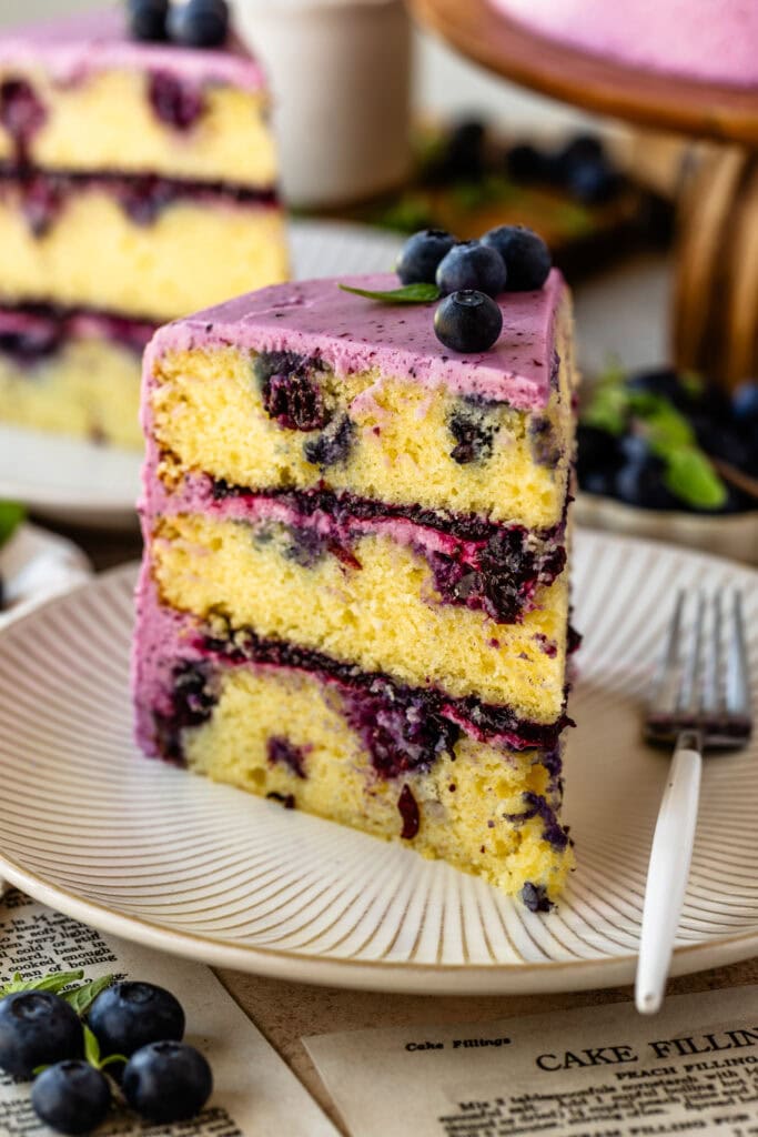 slice of blueberry cake on a plate loaded with blueberries, topped with frosting and blueberries on top.