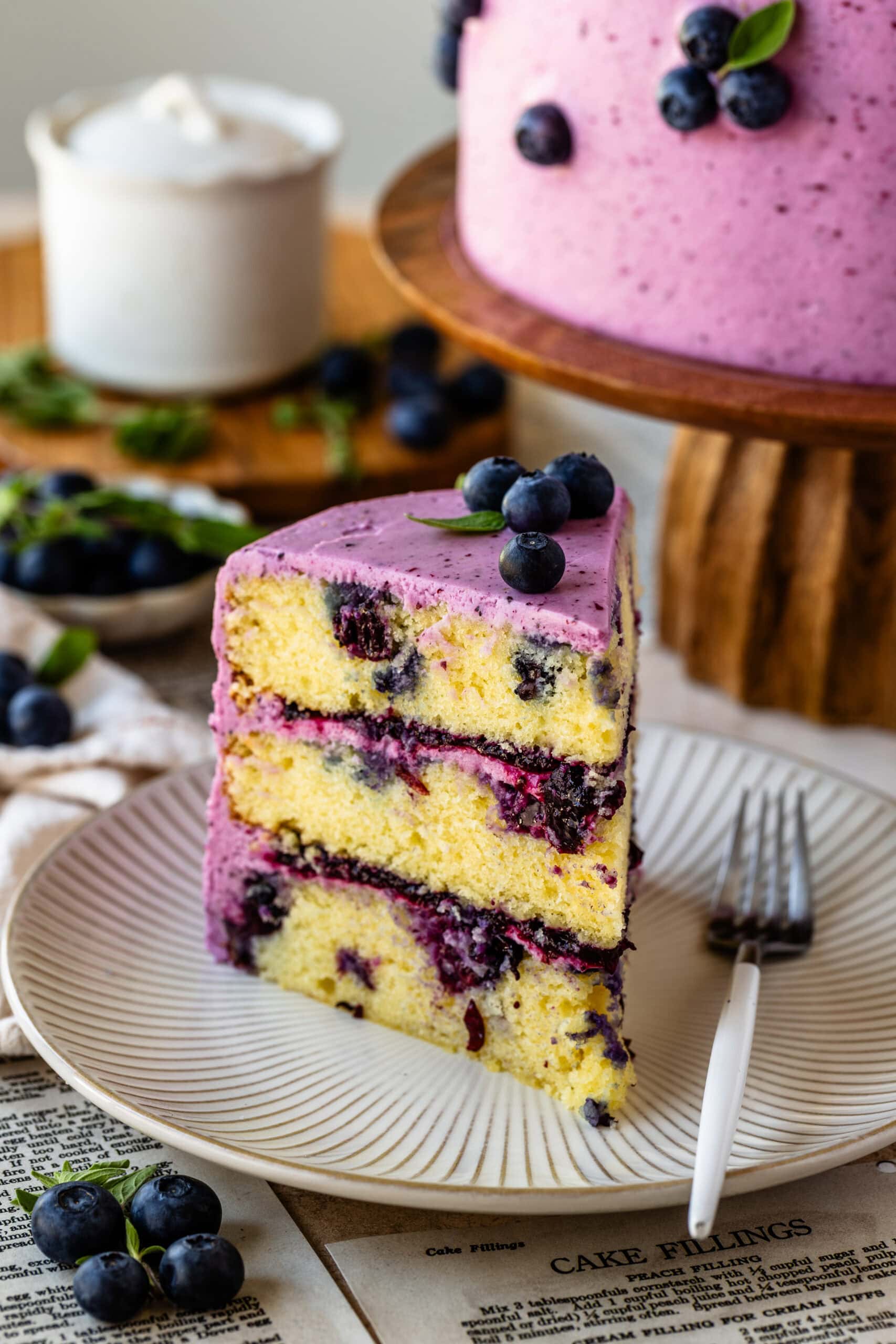 slice of blueberry cake on a plate loaded with blueberries, topped with frosting and blueberries on top.