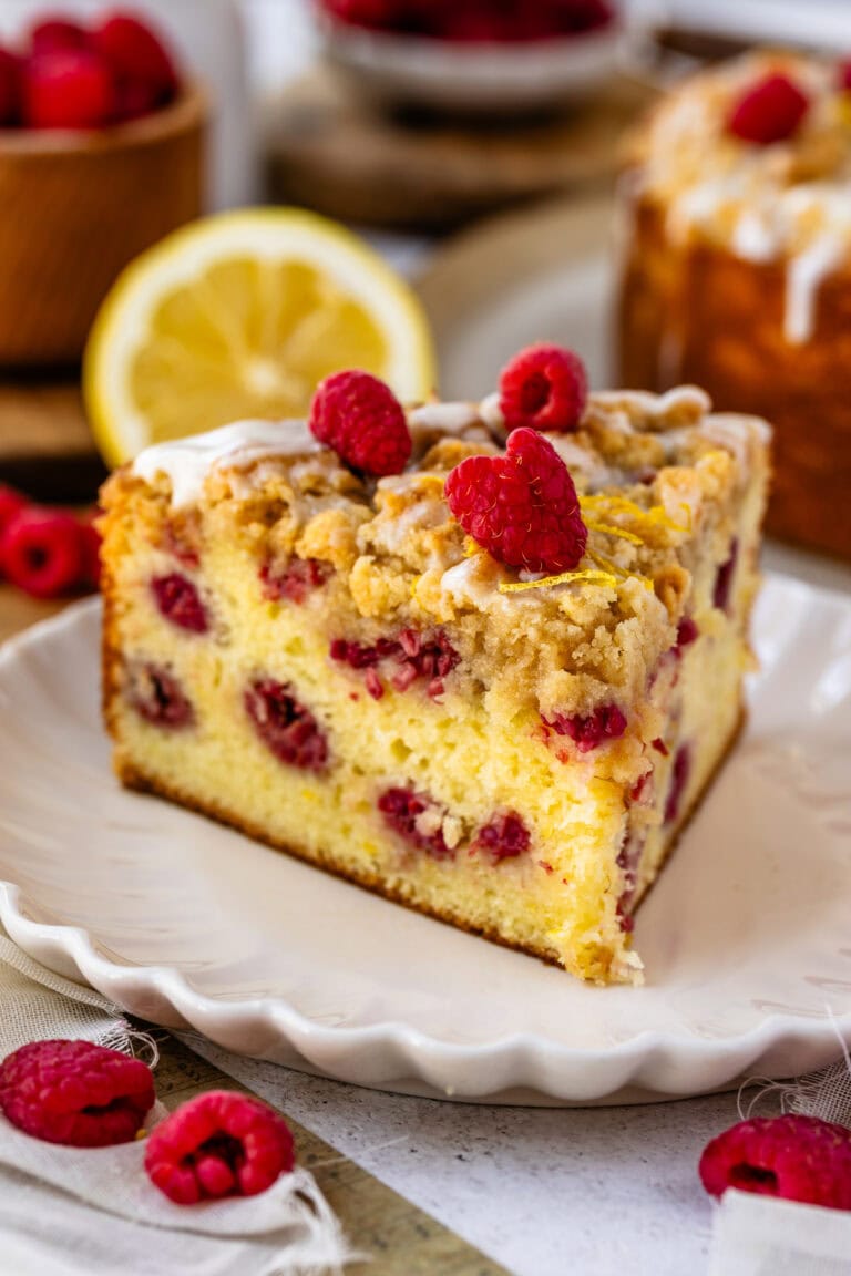 slice of lemon raspberry cake with glaze and crumble on top, and raspberries on top of the cake slice.