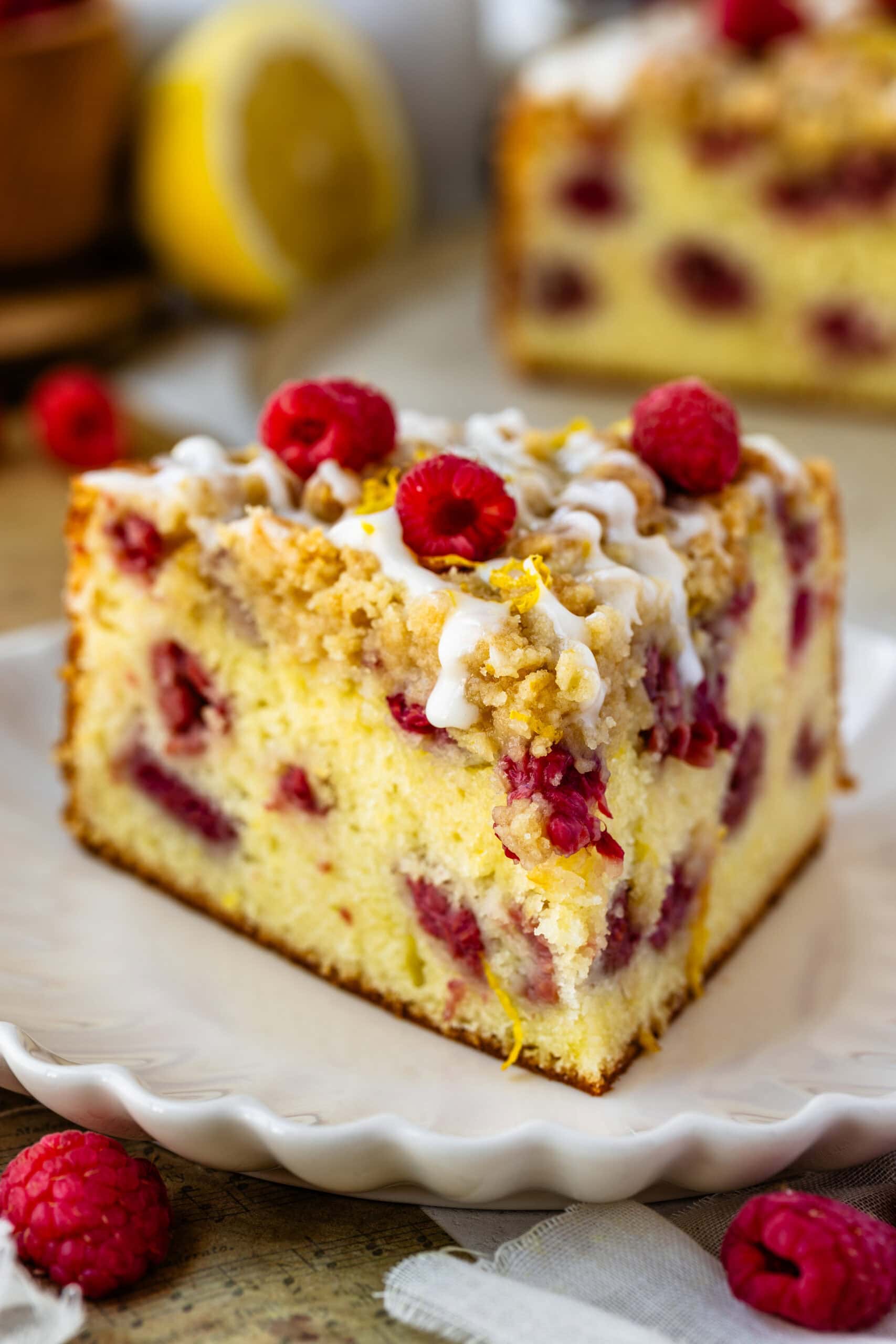 slice of lemon raspberry cake with glaze and crumble on top, and raspberries on top of the cake slice.