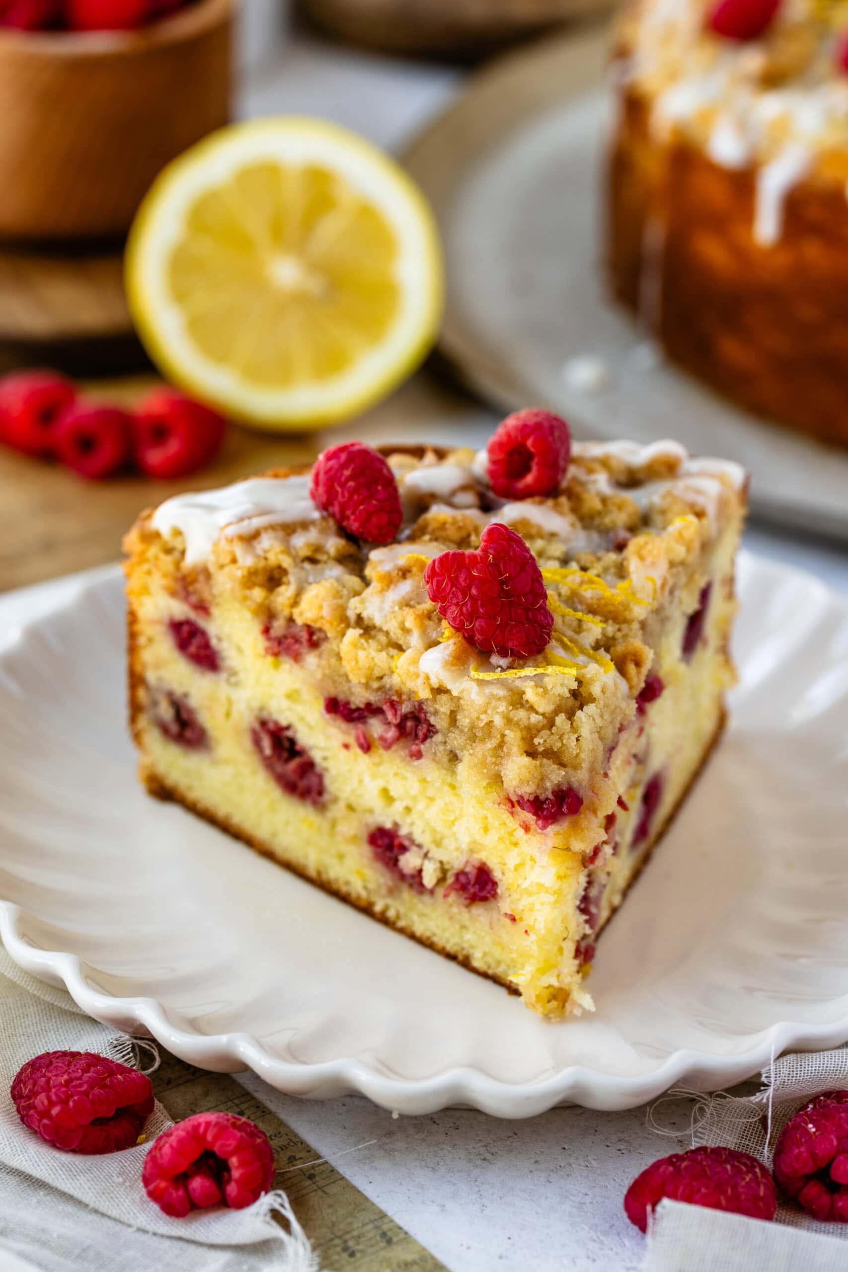slice of lemon raspberry cake with glaze and crumble on top, and raspberries on top of the cake slice.