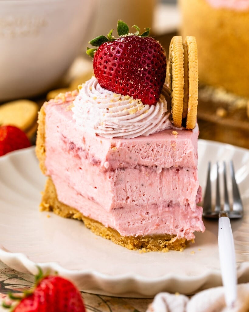 slice of no bake strawberry cheesecake on a plate topped with strawberry whipped cream, and fresh strawberry.