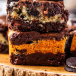 two oreo brownies stacked on top of each other, there's a layer of cookies and cream white brownie in between two layers of dark chocolate brownie in each slice. the slices are on top of a wooden board, with oreos around.