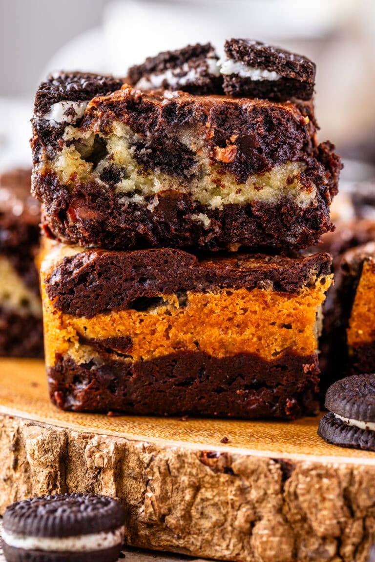 two oreo brownies stacked on top of each other, there's a layer of cookies and cream white brownie in between two layers of dark chocolate brownie in each slice. the slices are on top of a wooden board, with oreos around.
