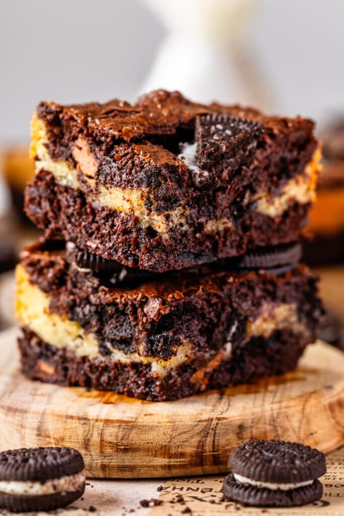two oreo brownies stacked on top of each other, there's a layer of cookies and cream white brownie in between two layers of dark chocolate brownie in each slice. the slices are on top of a wooden board, with oreos around.