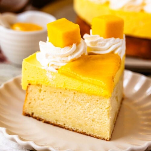 Mango Mousse Cake slice on a plate decorated with mango slices and whipped cream.