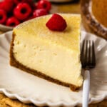 slice of new york cheesecake with powdered sugar on top and a raspberry, sitting on a plate with a fork on the plate.