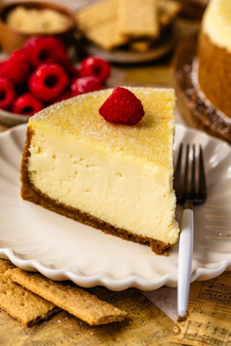 slice of new york cheesecake with powdered sugar on top and a raspberry, sitting on a plate with a fork on the plate.