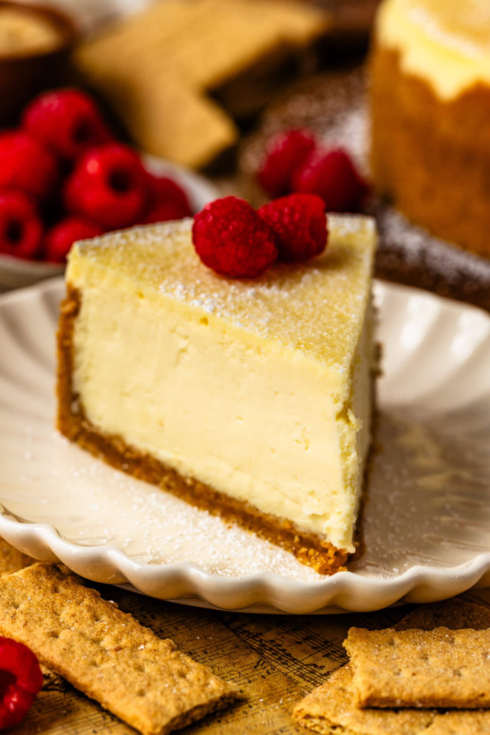 slice of new york cheesecake with powdered sugar on top and a raspberry, sitting on a plate with a fork on the plate.