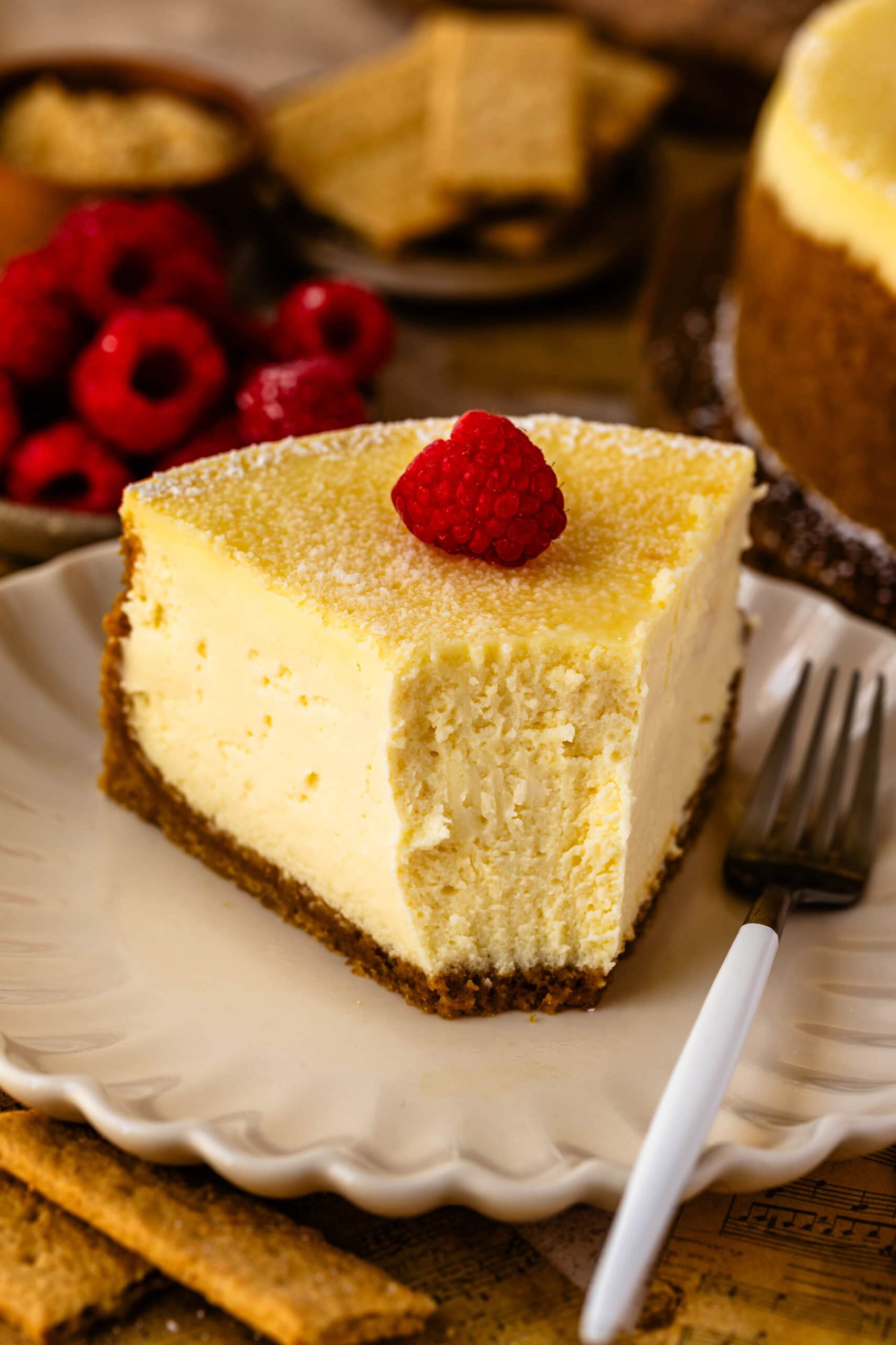 slice of new york cheesecake with powdered sugar on top and a raspberry, sitting on a plate with a fork on the plate.