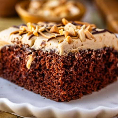 Chocolate Cake with Peanut Butter Frosting: A slice of rich, moist chocolate cake topped with a thick layer of peanut butter frosting. The frosting is garnished with chopped peanuts and a drizzle of chocolate. The cake slice is presented on a scalloped white plate, with the background showing a blurred rustic kitchen setting, including a bowl of peanuts.