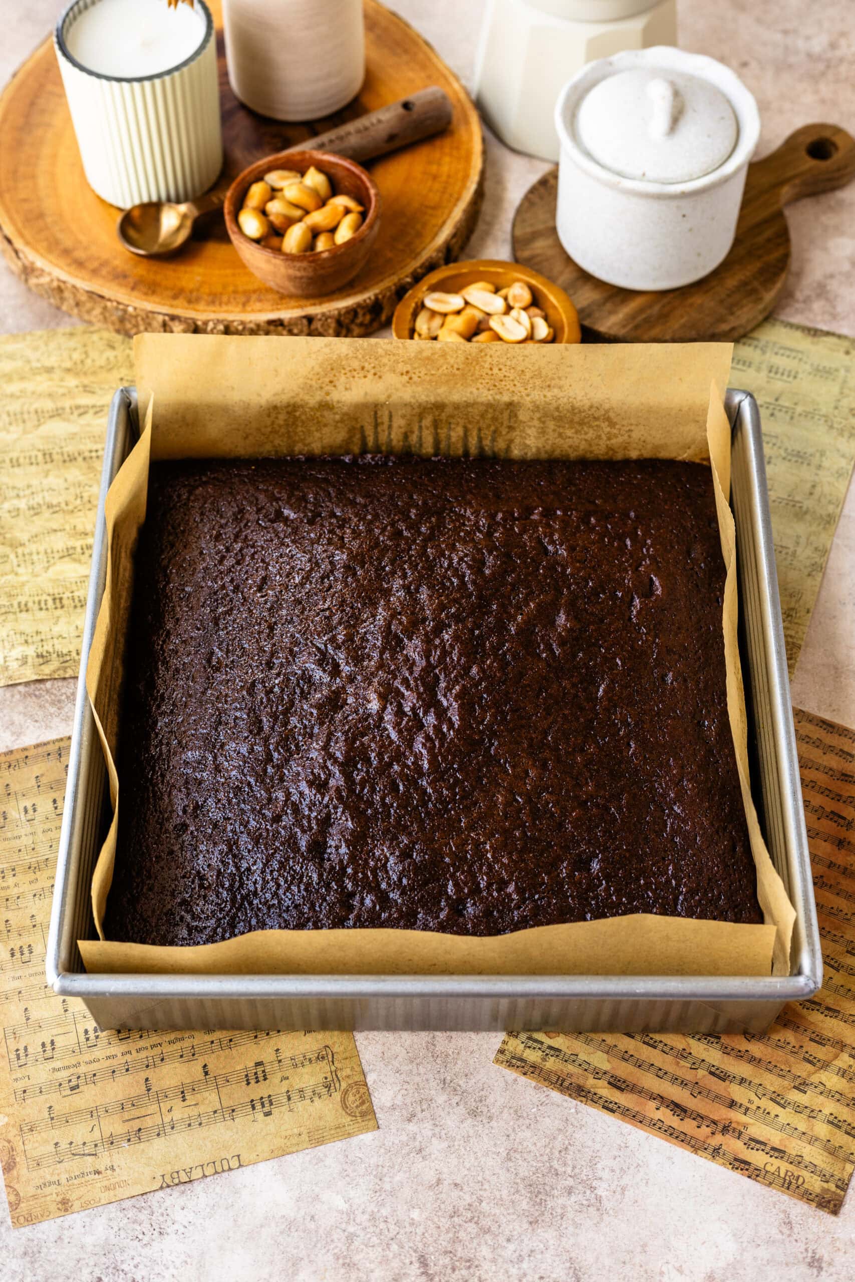 Baked Chocolate Cake in a Pan: A freshly baked chocolate cake in a square baking pan lined with parchment paper. The top of the cake has a rich, moist texture. The pan is set on a countertop, with a wooden tray holding candles, bowls of peanuts, and other kitchen items in the background.