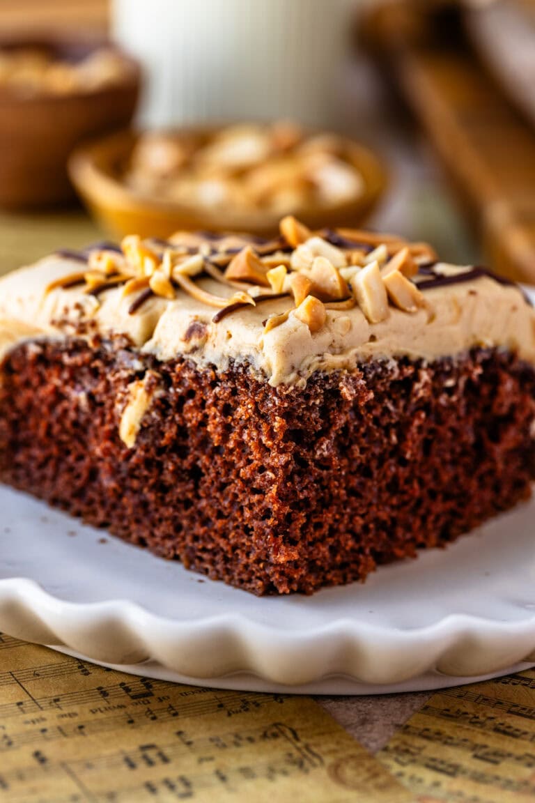 Chocolate Cake with Peanut Butter Frosting: A slice of rich, moist chocolate cake topped with a thick layer of peanut butter frosting. The frosting is garnished with chopped peanuts and a drizzle of chocolate. The cake slice is presented on a scalloped white plate, with the background showing a blurred rustic kitchen setting, including a bowl of peanuts.