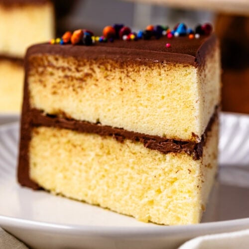 A close-up shot of a slice of two-layer yellow cake with rich chocolate frosting. The cake has a soft, fluffy texture, and the chocolate frosting on top is smooth with colorful sprinkles for decoration. The slice is served on a white plate with a soft cloth napkin in the background, giving the image a cozy and inviting feel.