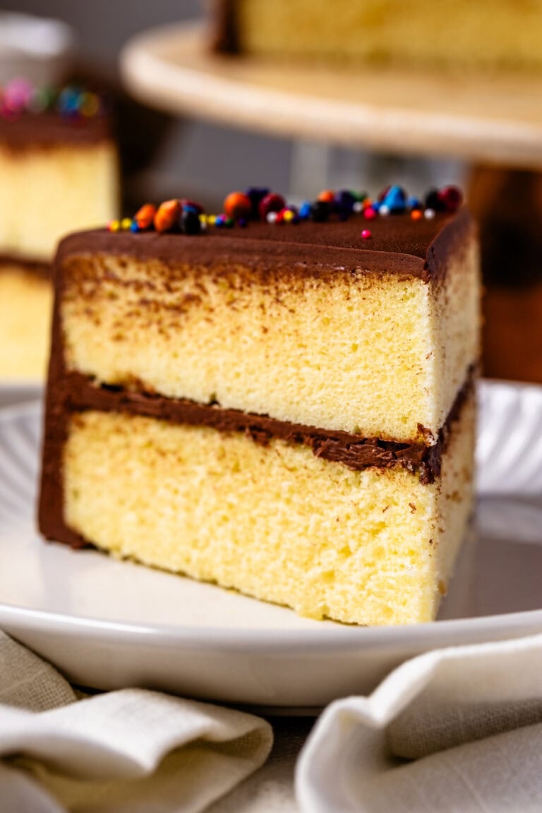 A close-up shot of a slice of two-layer yellow cake with rich chocolate frosting. The cake has a soft, fluffy texture, and the chocolate frosting on top is smooth with colorful sprinkles for decoration. The slice is served on a white plate with a soft cloth napkin in the background, giving the image a cozy and inviting feel.
