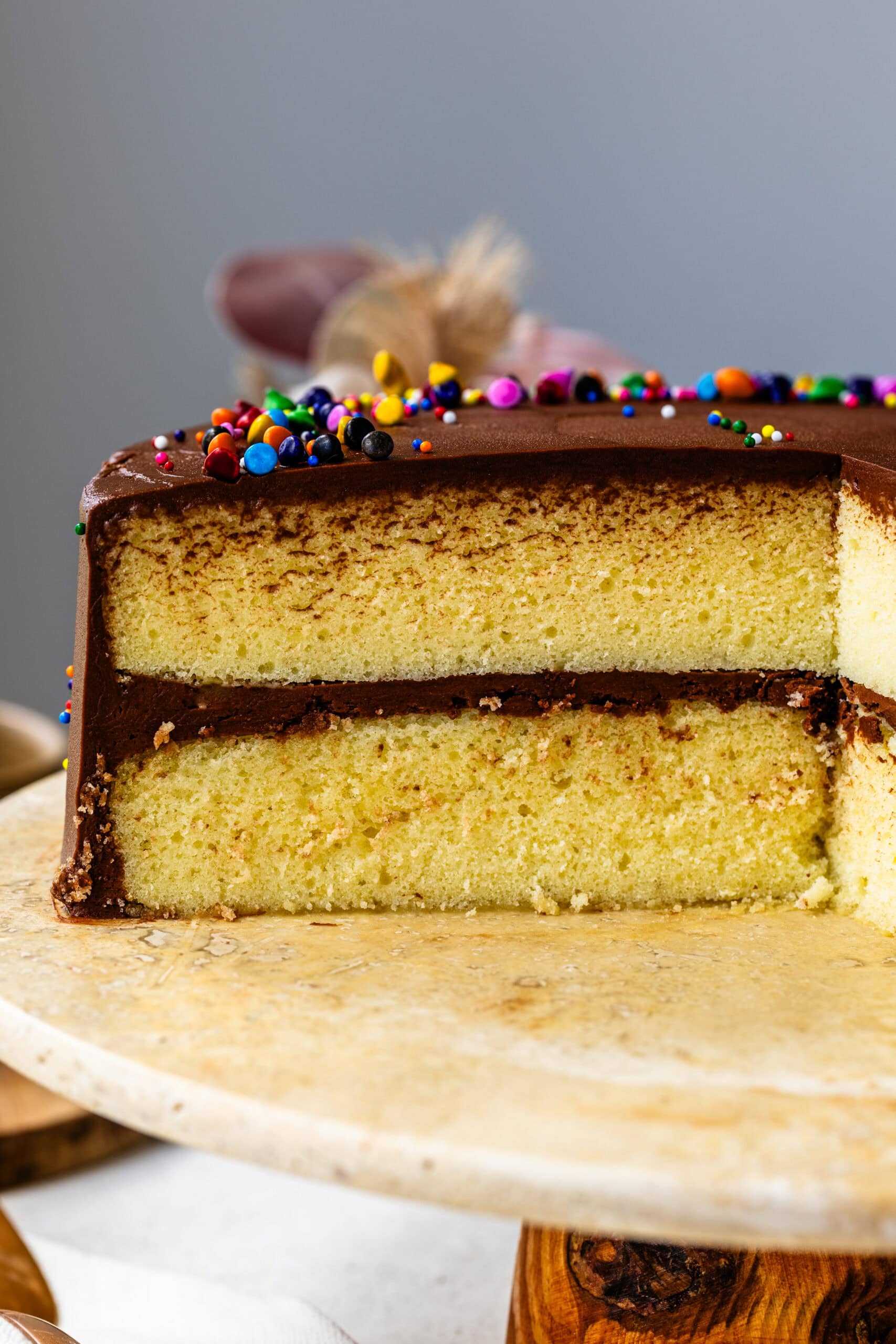 A beautifully frosted yellow cake with a smooth layer of chocolate buttercream and colorful sprinkles on top. A slice of the cake has been removed, revealing two moist, fluffy yellow cake layers with a thick chocolate frosting in between. The cake sits on a rustic stone cake stand with a soft cloth draped around the base, creating a cozy and inviting presentation. In the background, there are soft floral decorations that add a touch of elegance to the scene.