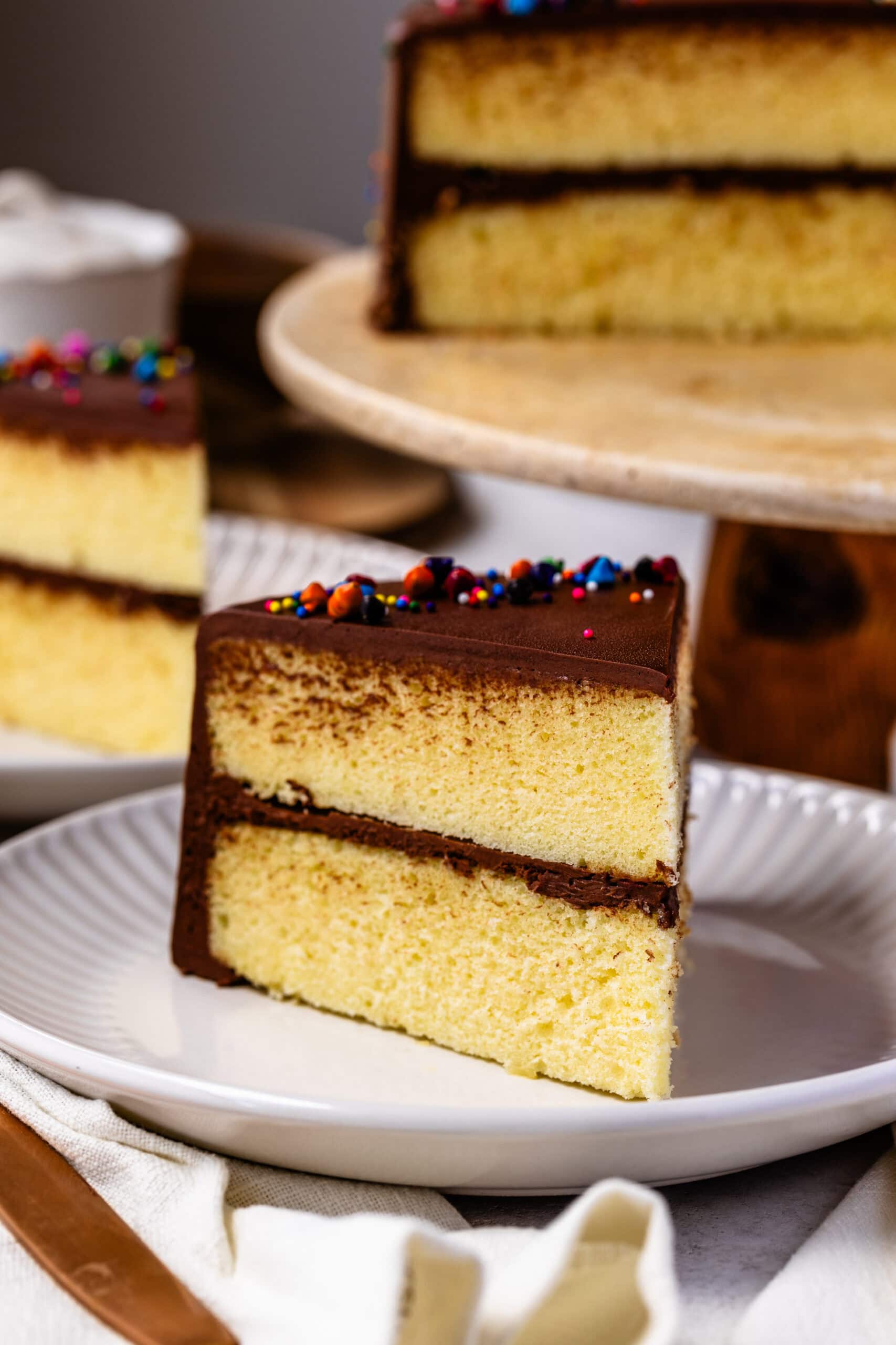 A close-up shot of a slice of two-layer yellow cake with rich chocolate frosting. The cake has a soft, fluffy texture, and the chocolate frosting on top is smooth with colorful sprinkles for decoration. The slice is served on a white plate with a soft cloth napkin in the background, giving the image a cozy and inviting feel.