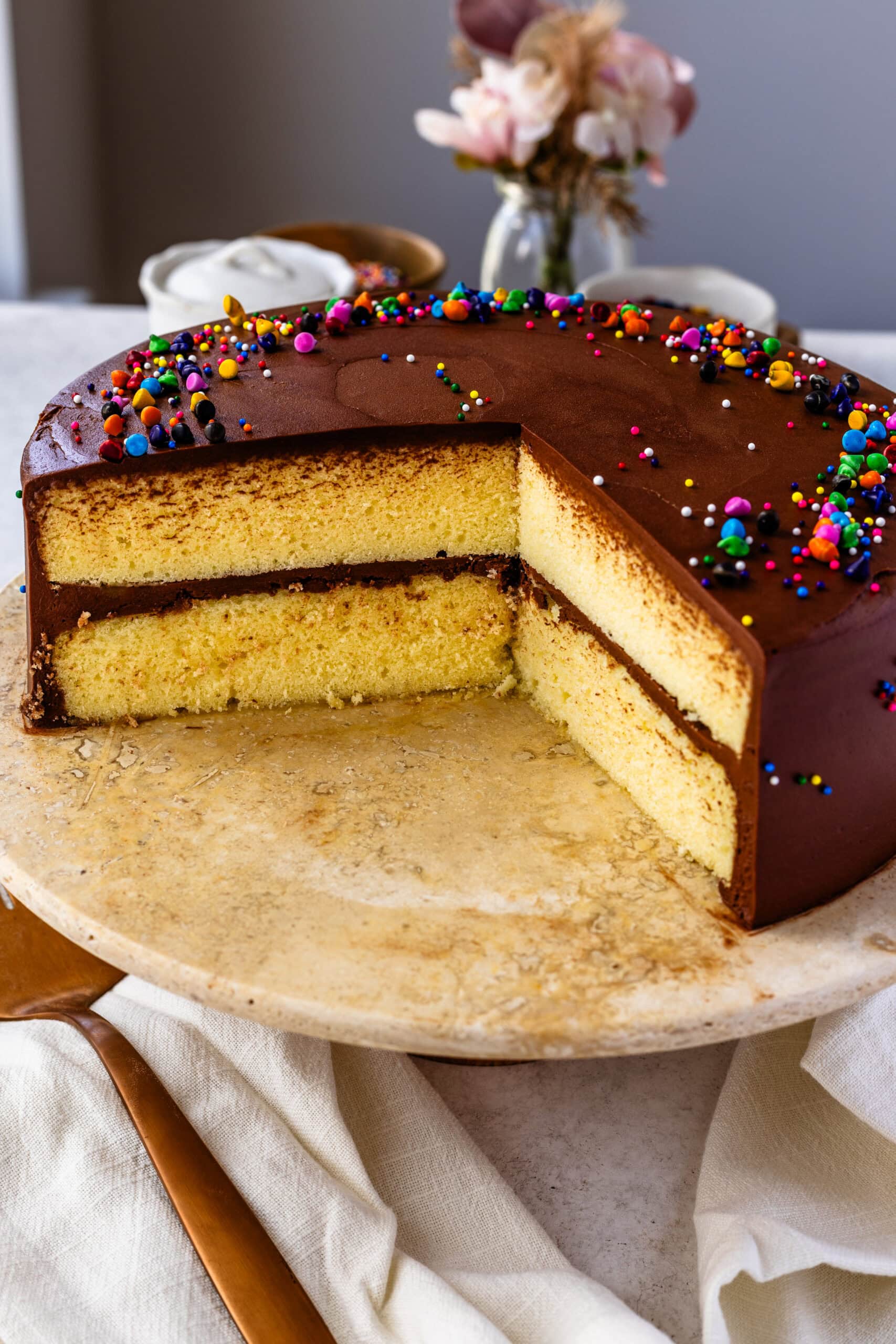 A beautifully frosted yellow cake with a smooth layer of chocolate buttercream and colorful sprinkles on top. A slice of the cake has been removed, revealing two moist, fluffy yellow cake layers with a thick chocolate frosting in between. The cake sits on a rustic stone cake stand with a soft cloth draped around the base, creating a cozy and inviting presentation. In the background, there are soft floral decorations that add a touch of elegance to the scene.