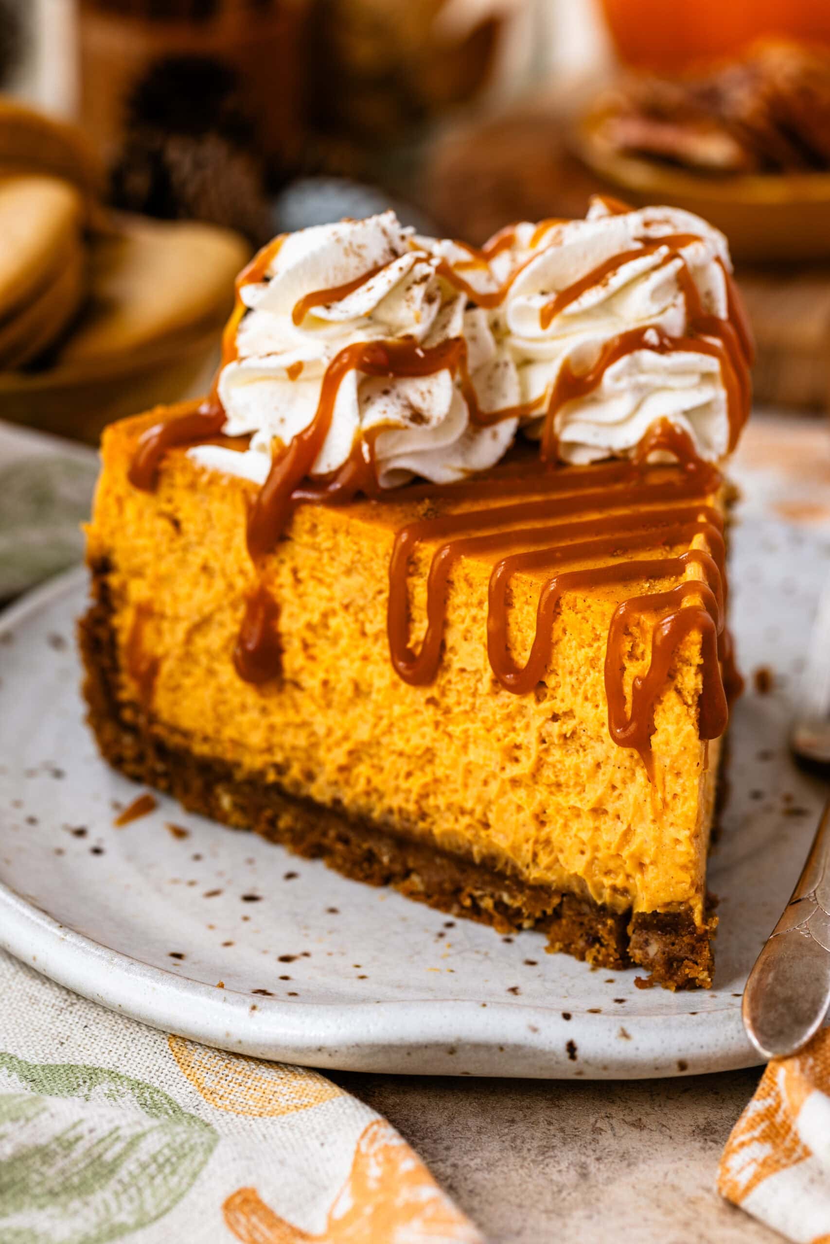 A slice of pumpkin cheesecake topped with whipped cream rosettes and drizzled with caramel sauce, placed on a ceramic plate. The creamy pumpkin filling and graham cracker crust are highlighted, with a cozy fall-themed background featuring cookies, nuts, and autumn decorations.