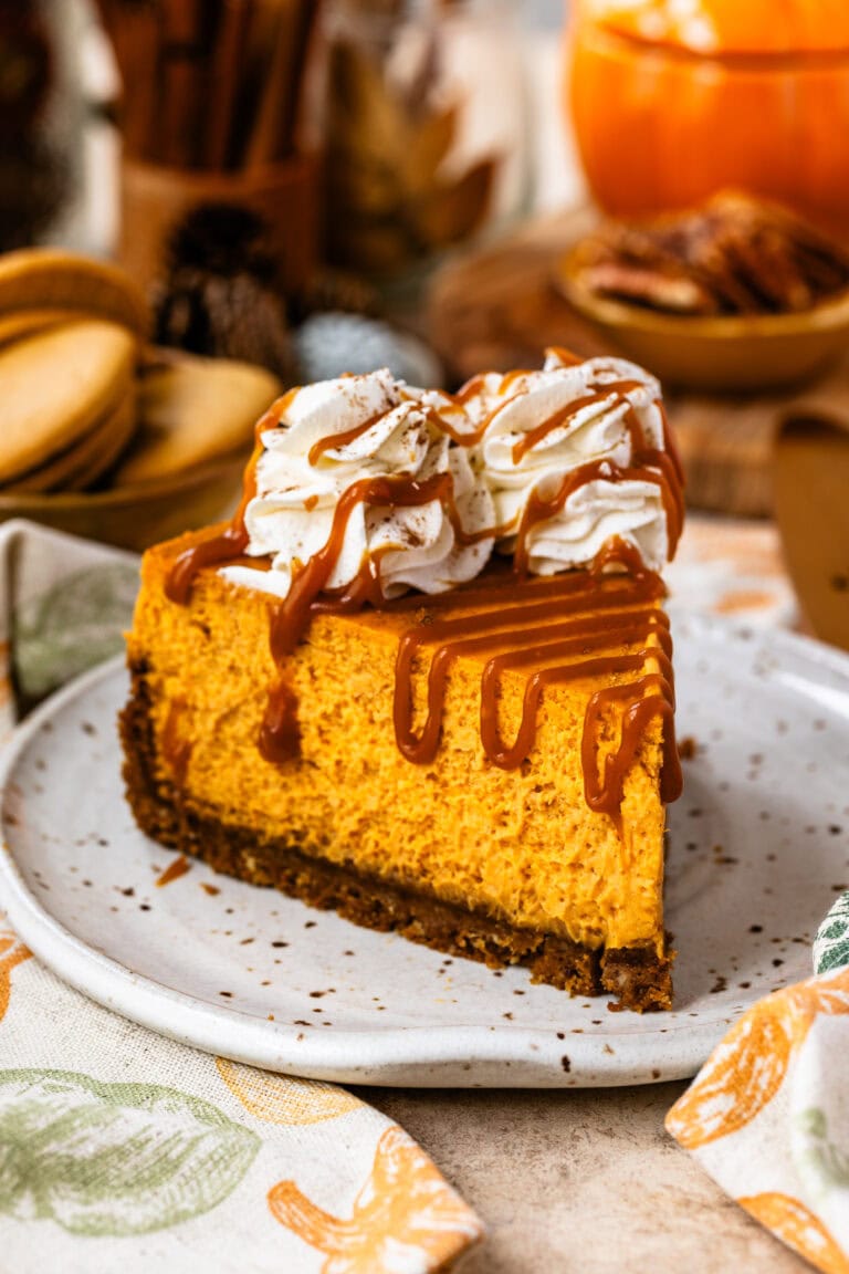 A slice of pumpkin cheesecake topped with whipped cream rosettes and drizzled with caramel sauce, placed on a ceramic plate. The creamy pumpkin filling and graham cracker crust are highlighted, with a cozy fall-themed background featuring cookies, nuts, and autumn decorations.