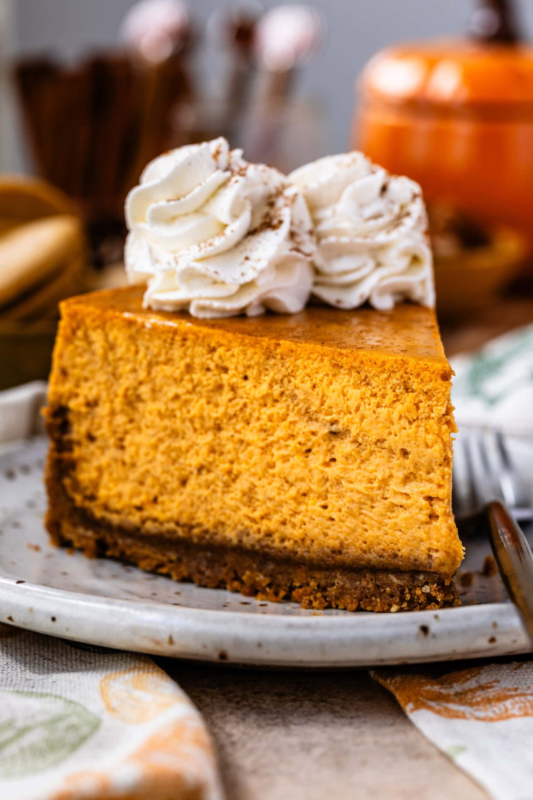 A slice of pumpkin cheesecake on a plate, topped with whipped cream rosettes dusted with cinnamon, showing the creamy texture of the cheesecake with a graham cracker crust. In the background, there are cookies and a wooden board, creating a cozy autumn vibe.