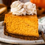 A slice of pumpkin cheesecake on a plate, topped with whipped cream rosettes dusted with cinnamon, showing the creamy texture of the cheesecake with a graham cracker crust. In the background, there are cookies and a wooden board, creating a cozy autumn vibe.