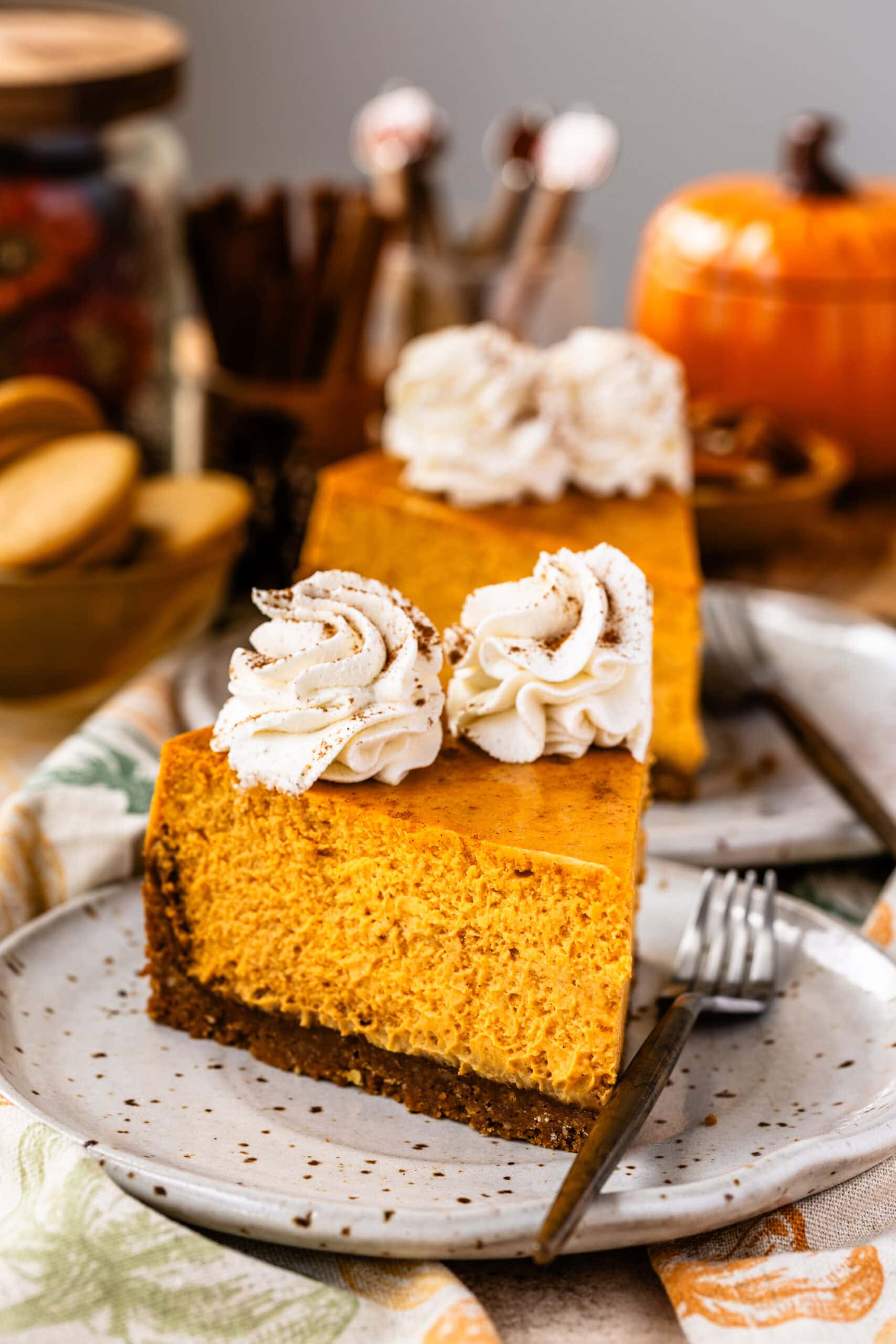 Two slices of pumpkin cheesecake topped with whipped cream rosettes and sprinkled with cinnamon, served on ceramic plates. The scene includes fall-themed decorations such as a ceramic pumpkin, cookies, and a warm, rustic atmosphere.