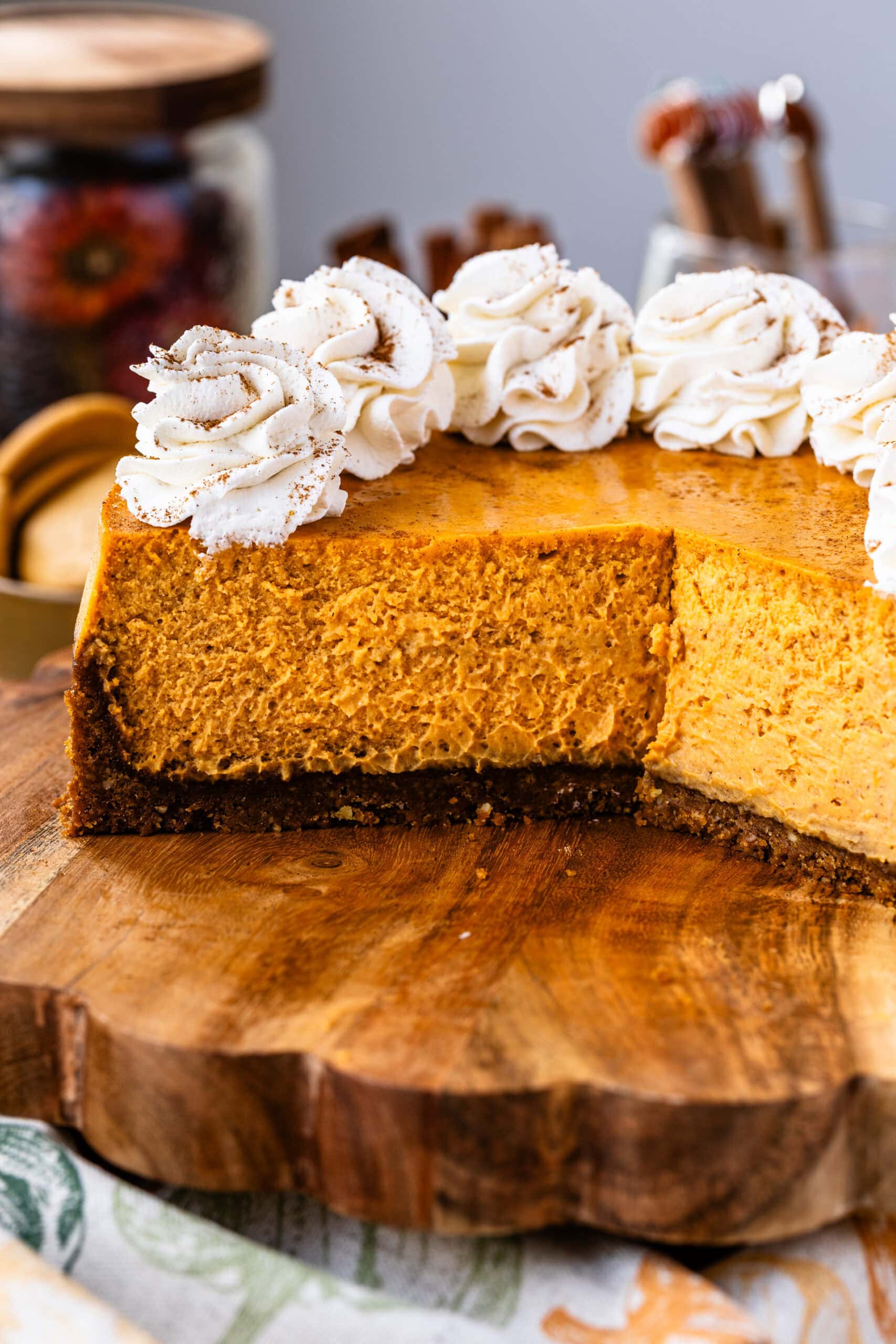A close-up of a pumpkin cheesecake with a slice cut out, showing the rich and fluffy texture of the cheesecake. The remaining cheesecake is decorated with whipped cream rosettes and sprinkled with cinnamon, placed on a rustic wooden board with fall decorations in the background.